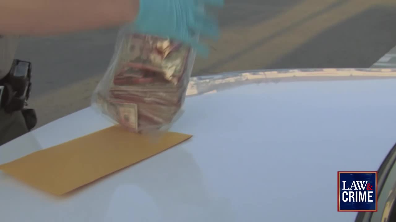 A gloved hand is lowering a clear plastic bag filled with stacks of cash onto the hood of a car. Below the bag, a yellow envelope sits on the white surface.

