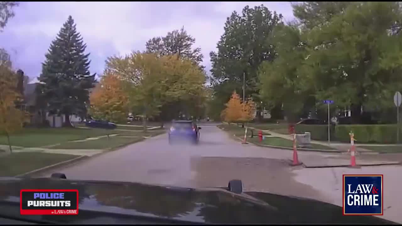 A blue car, likely in pursuit, is speeding down a wet residential street. The camera is mounted on a police vehicle, as indicated by the "Police Pursuits" logo from Law & Crime.
