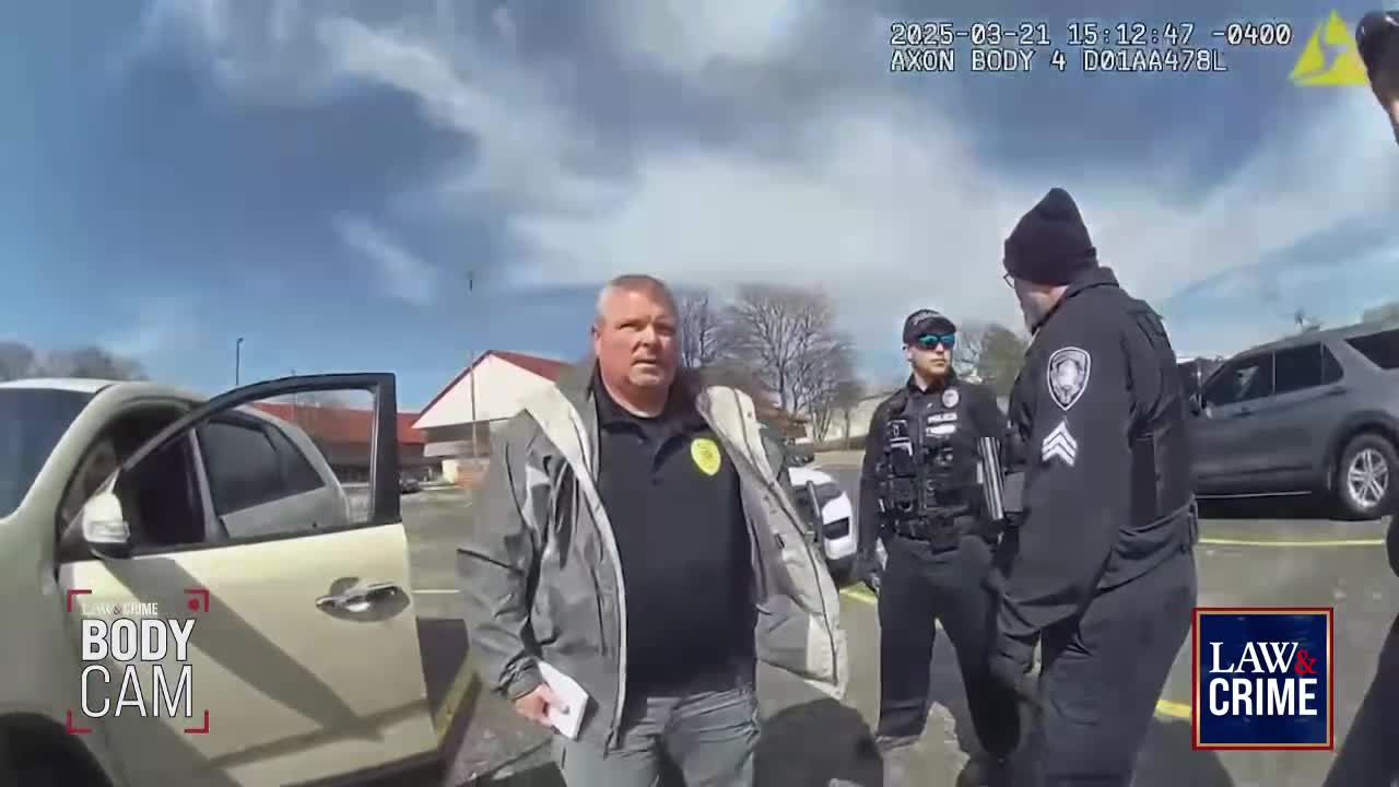 A man in a grey jacket stands near a car with its door open, facing the camera. Two police officers in uniform are positioned to his right, engaged in conversation.
