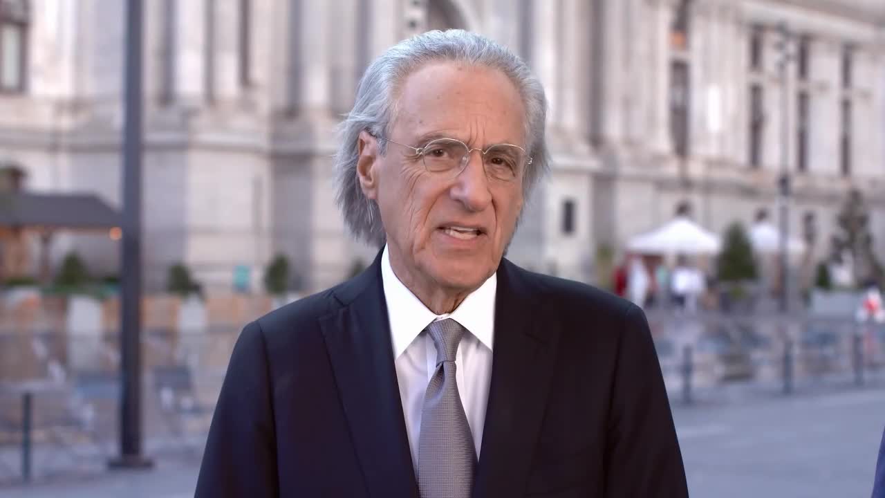 A man in a suit and tie is speaking, framed against a blurred background of a building in the United States. He's likely delivering a report for Law & Crime.
