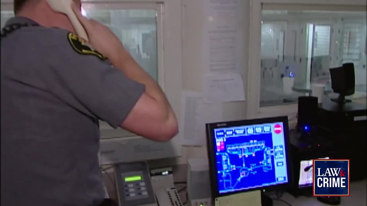 A uniformed officer holds a phone to his ear, his back to the camera. The screen in front of him displays a blue map, likely surveillance footage from inside a facility.
