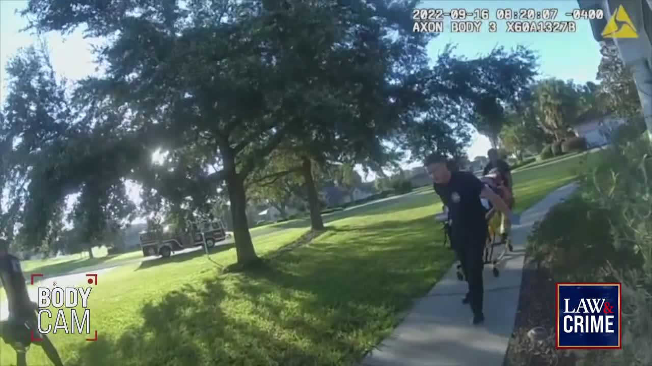 A police officer is pushing a gurney down a sidewalk, another person walking behind. The sun is bright, and the scene is framed by trees and a grassy lawn.
