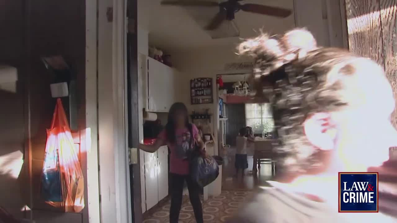 A young person, holding a bag, stands in a doorway, facing the camera. The camera is positioned close to another person's head, partially obscuring the view.
