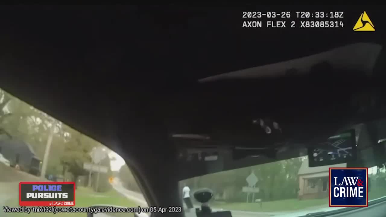 The camera is mounted inside a vehicle, likely a police car, as it navigates a residential street. A person is visible on the side of the road, and the car is moving forward.
