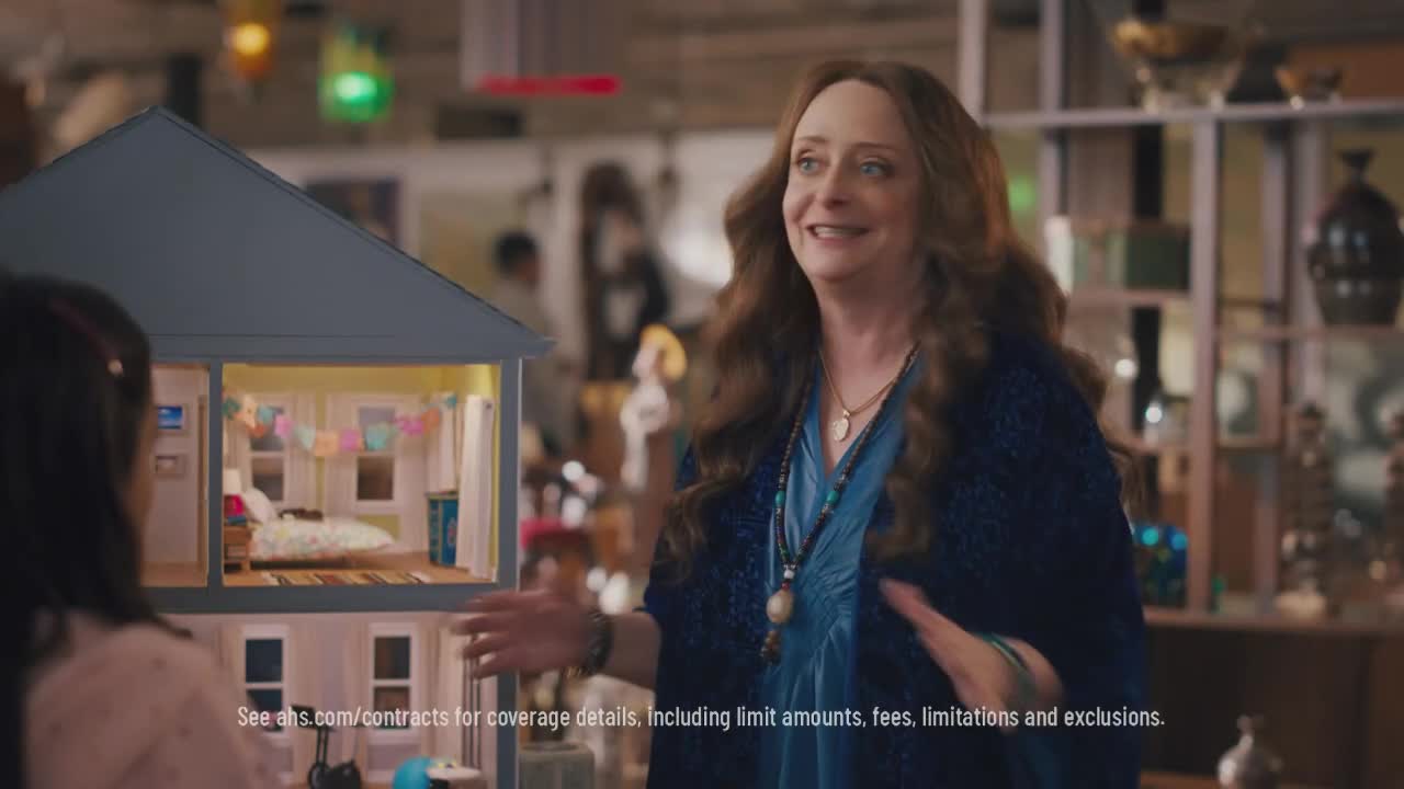 A woman with long, auburn hair gestures toward a dollhouse, smiling at someone off-camera. The dollhouse's interior is lit, revealing a miniature bedroom with a bed and colorful decorations.
