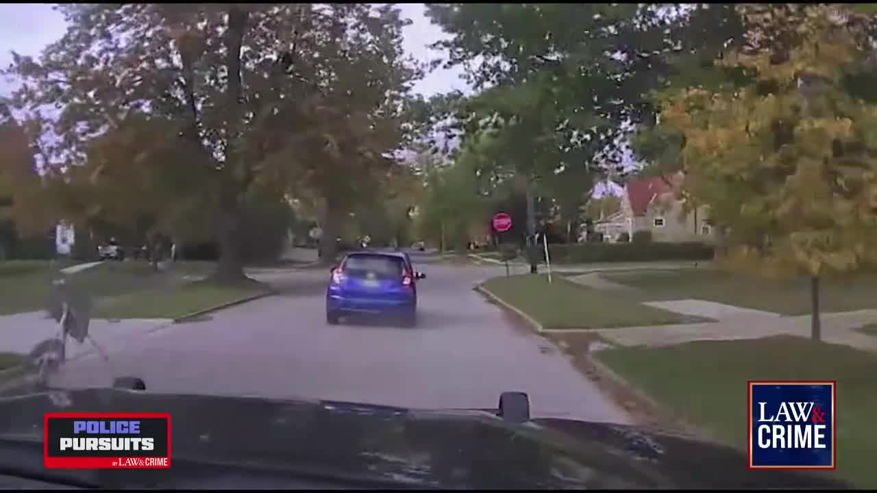 A blue car is moving down a residential street, seen from the perspective of a pursuing police vehicle. The Law & Crime logo is visible in the corner.
