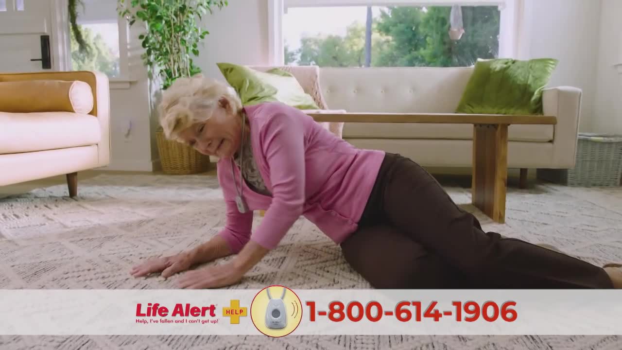 An elderly woman, wearing a pink sweater and brown pants, is on the floor, leaning on her hands. She appears to be struggling to get up, with a Life Alert pendant around her neck.
