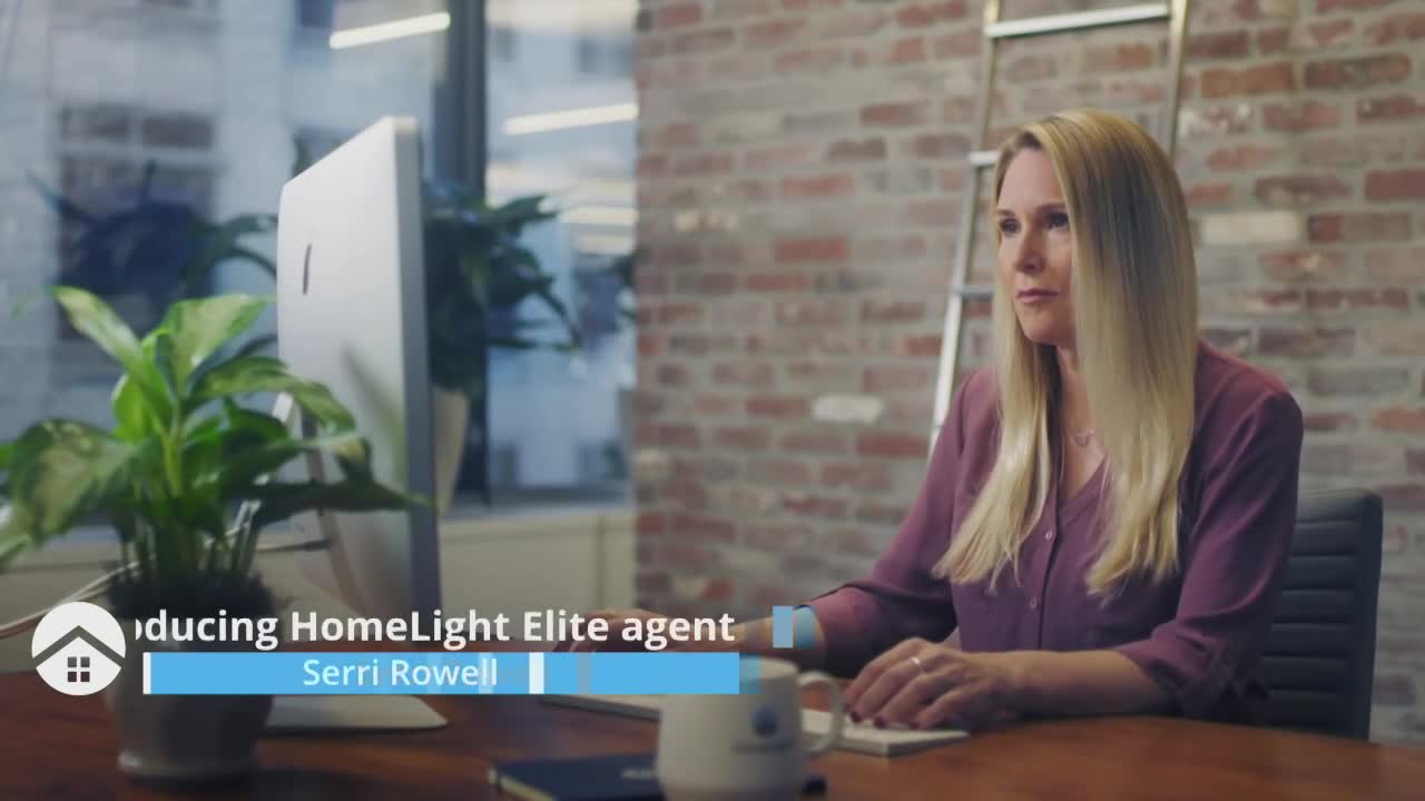Serri Rowell, a HomeLight Elite agent, sits at a desk, typing on a white keyboard in front of a computer screen. She's in a modern office, with a brick wall and a potted plant nearby.
