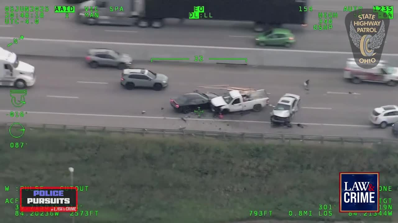 A mangled black car and a white pickup truck are smashed together on the highway. Debris litters the road as a person stands near the wreckage.
