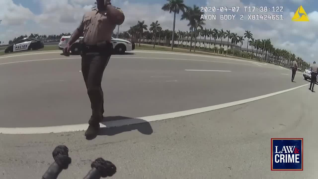 A Florida officer, phone to his ear, stands on the road's edge, gesturing with his left hand. Two patrol cars sit in the background, and other officers are visible further down the street.
