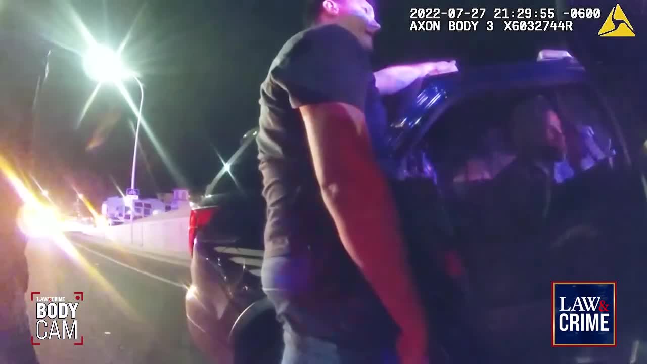 A man in a dark shirt leans into a car, illuminated by the bright streetlights. The scene is captured on a body camera, possibly during a Law & Crime investigation in the United States.

