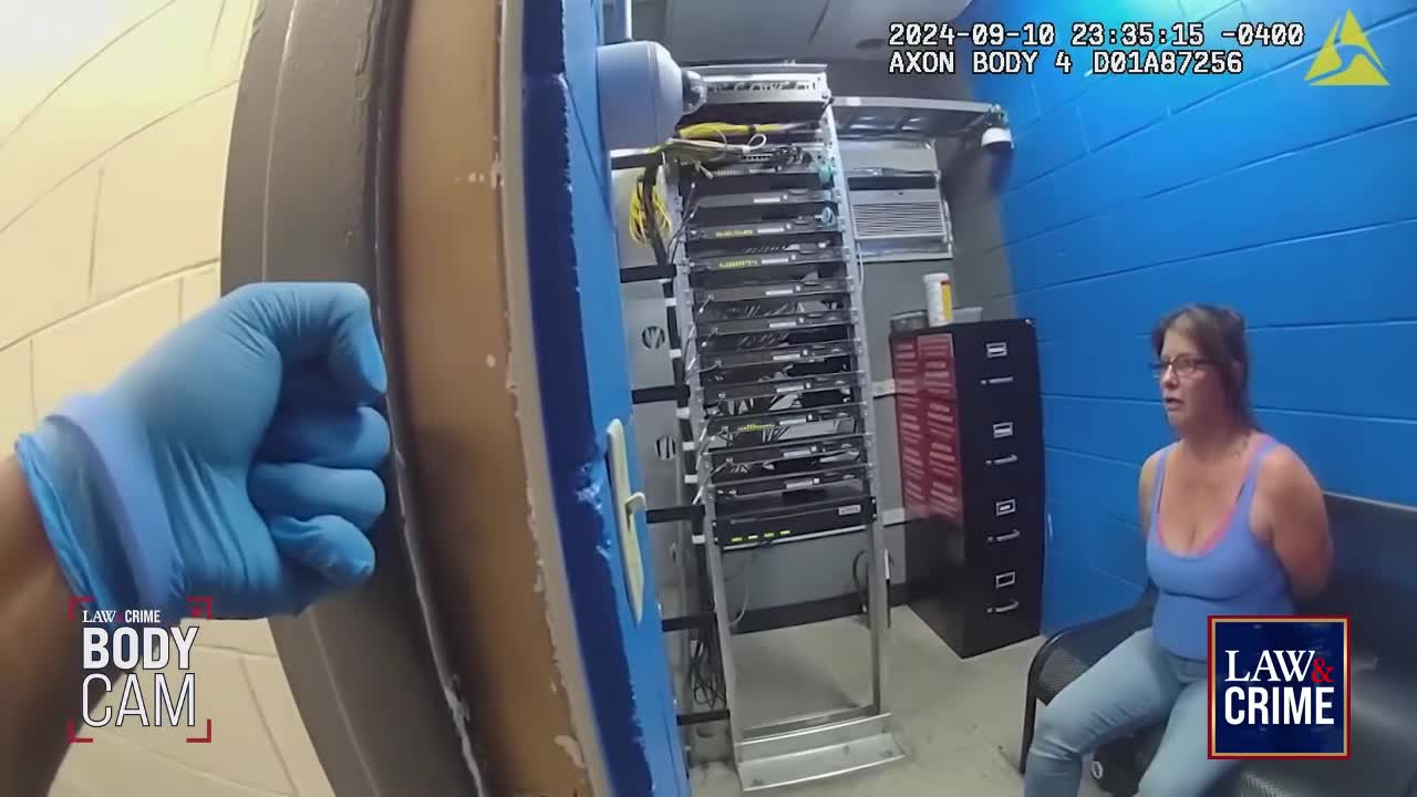 A gloved hand is pushing open a door, revealing a server rack and a woman seated on a bench, her hands cuffed behind her back. The woman in a blue tank top looks directly at the camera with a concerned expression.
