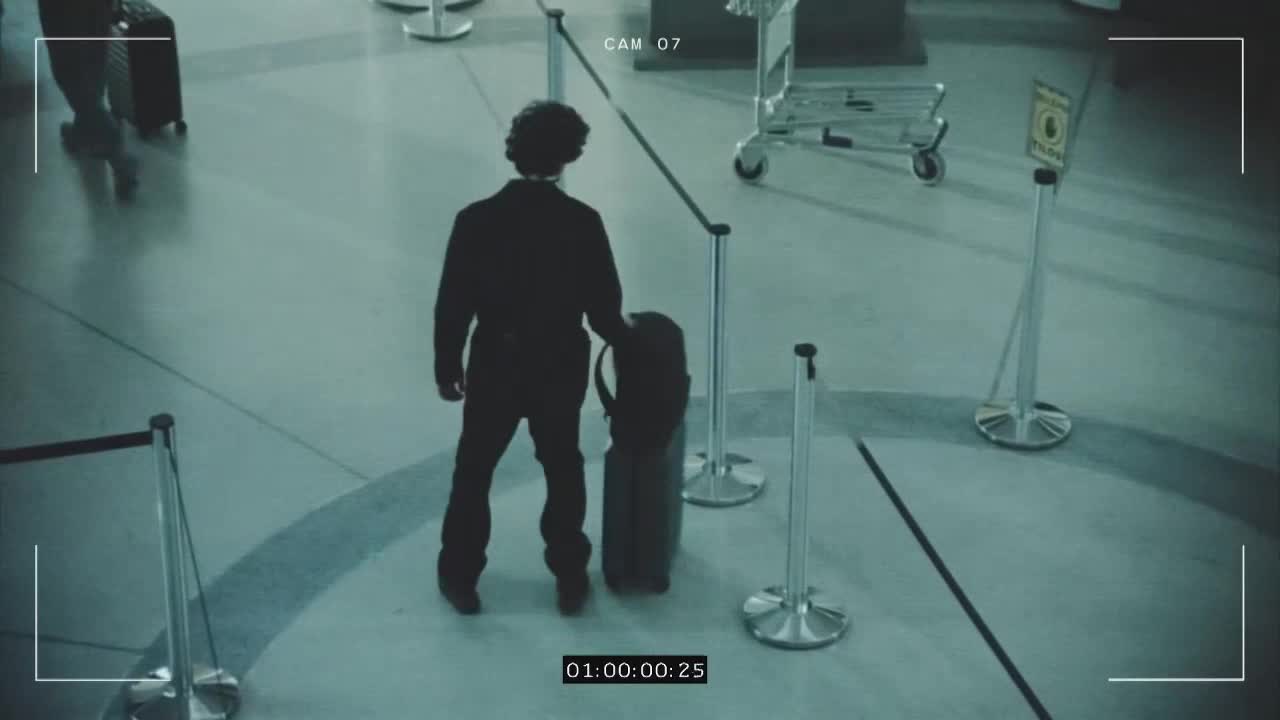 A man in a dark suit stands near a roped-off area, holding a large, black bag. He appears to be waiting, or perhaps considering his next move.
