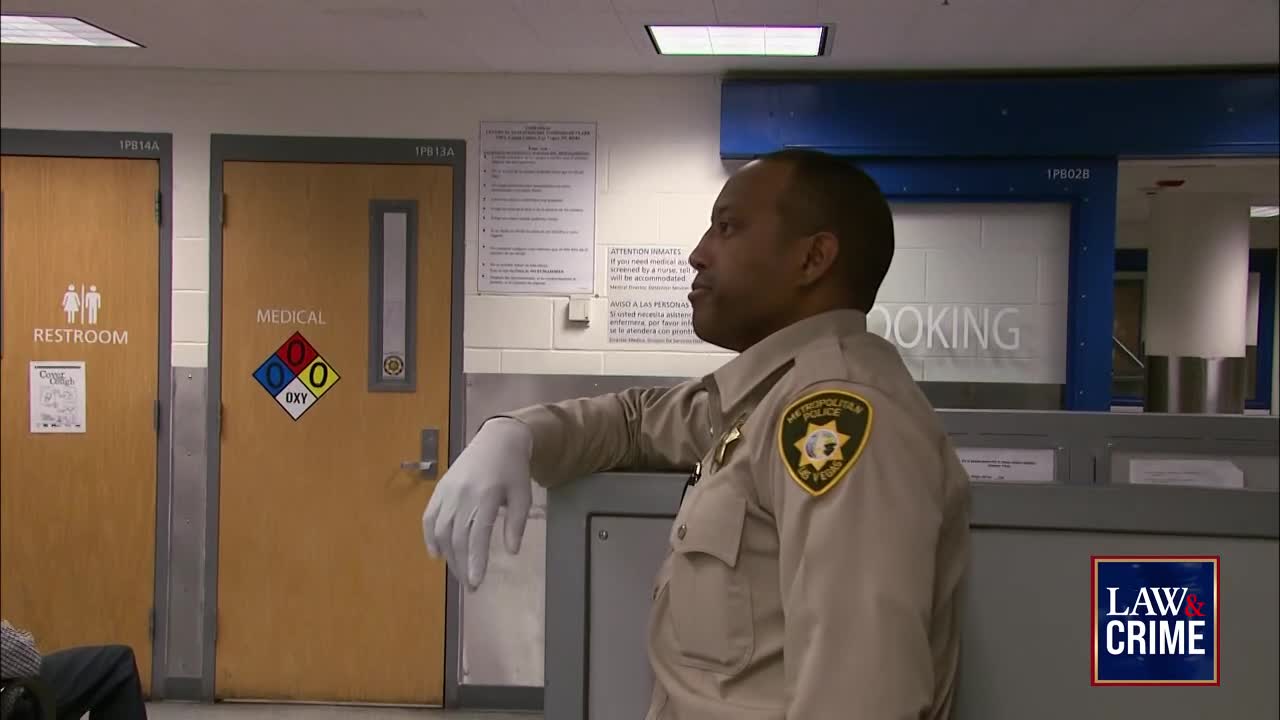 A uniformed officer, wearing gloves, leans against a counter in what appears to be a booking area. The "Law & Crime" logo is in the bottom right corner, and the officer is looking to the side.
