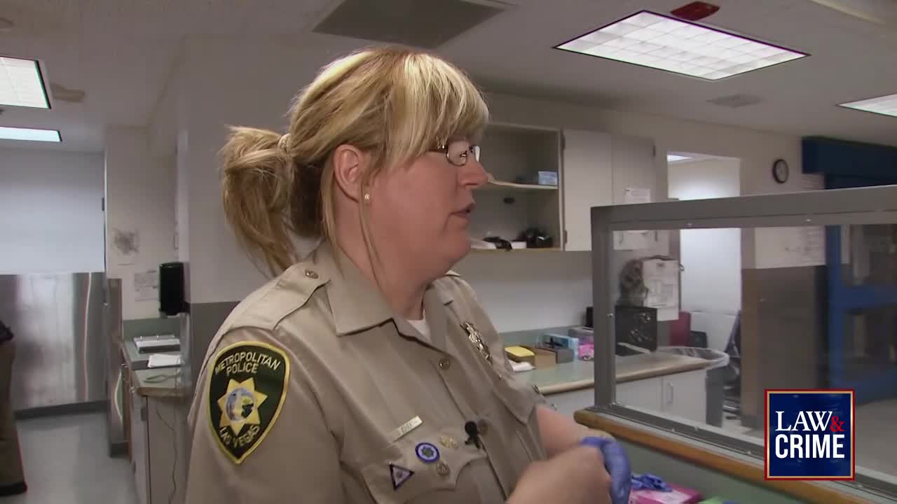A woman in a Metropolitan Police uniform is putting on blue gloves. She stands behind a clear partition, likely in a holding area.
