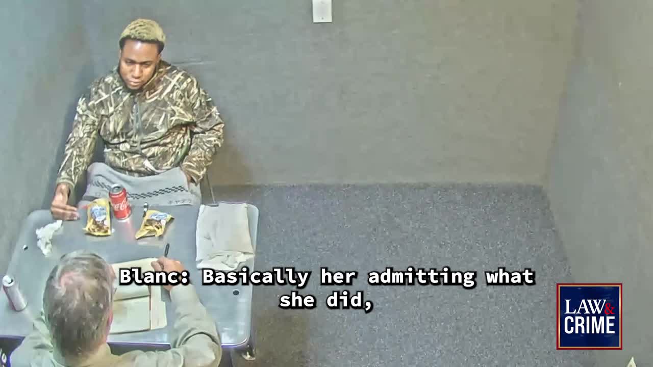 A man in a camouflage hoodie sits across a table from another man, who is writing in a notebook. A can of Coca-Cola and two bags of chips rest on the table between them.