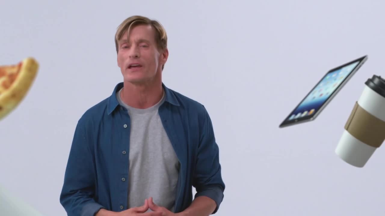 A slice of pizza drifts left, while a tablet and coffee cup float right. The man in the center, dressed in a blue shirt over a grey t-shirt, speaks with his hands clasped.