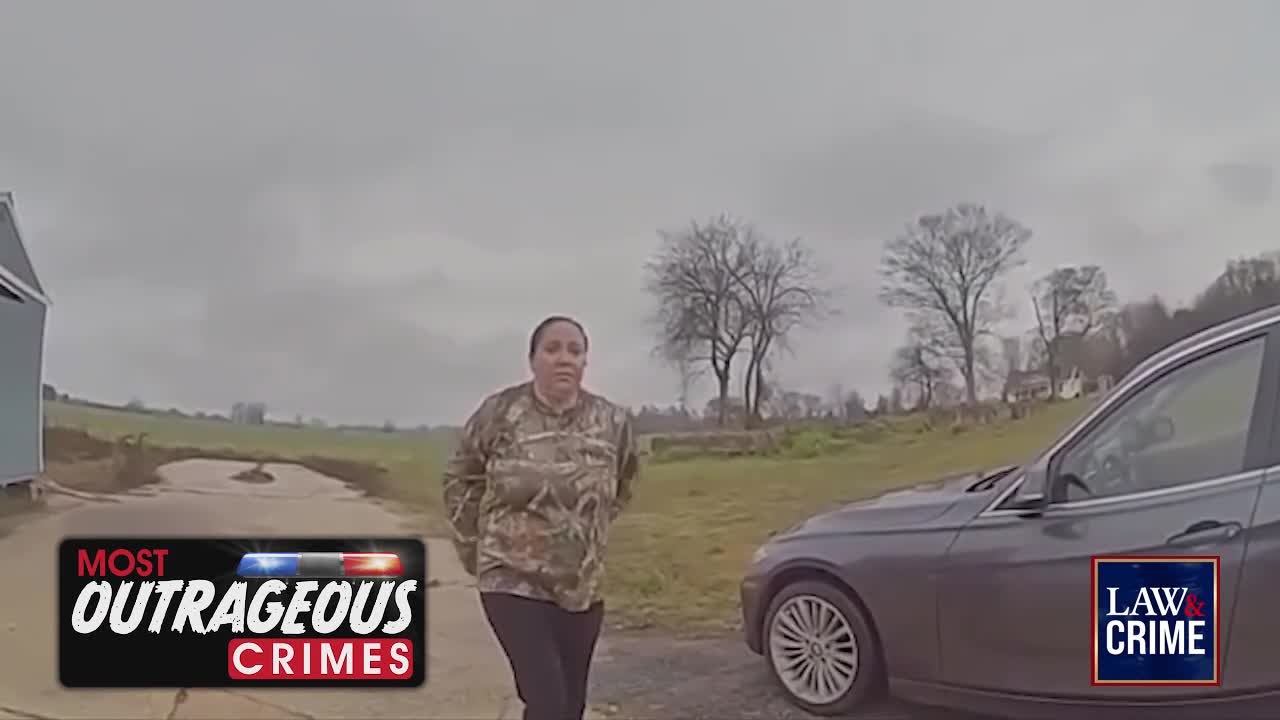 A woman in camouflage stands with her hands behind her back, facing a dark sedan parked on a gravel drive. The sky is overcast, and bare trees dot the landscape behind her.