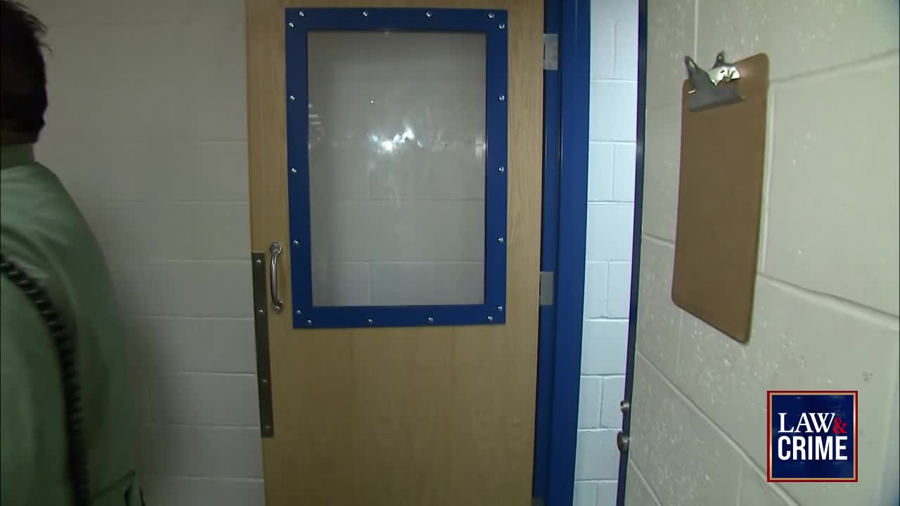 A man in a light green shirt walks away from a door with a small window. A clipboard hangs on the white cinder block wall to the right.