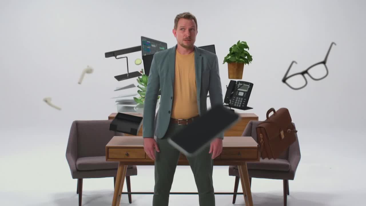 A man stands amidst a chaotic scene of office supplies that are suspended in mid-air around him. A tablet is falling directly in front of him, while a pair of glasses floats to his right.