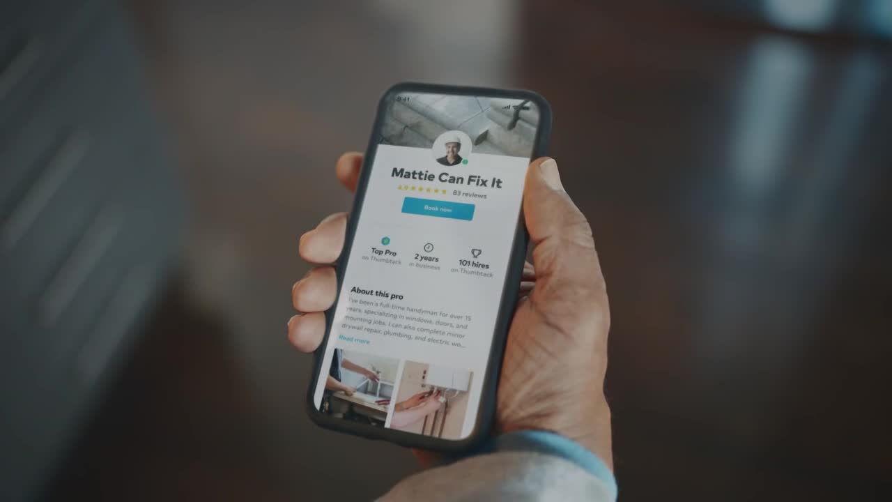 A hand holds a smartphone displaying a profile for "Mattie Can Fix It." The screen shows a "Book Now" button and details about the handyman's experience.