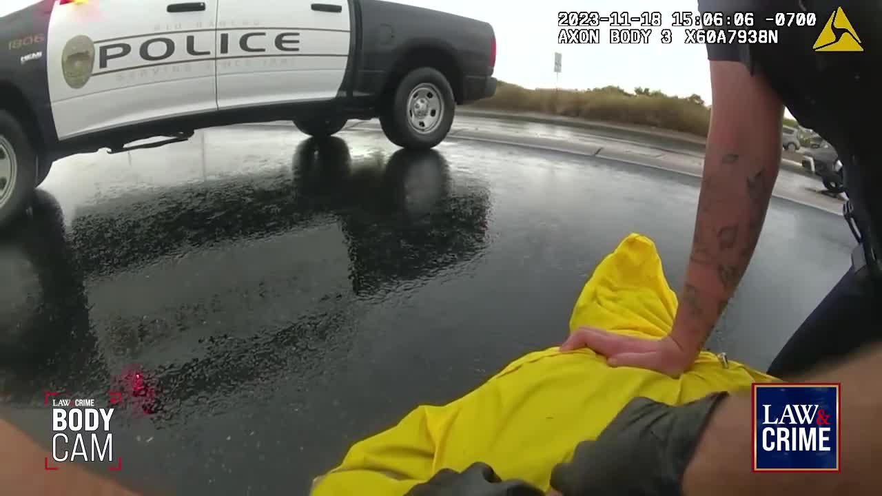A police officer is holding down a person in a bright yellow jacket on a wet road. A police vehicle sits nearby, its lights flashing.