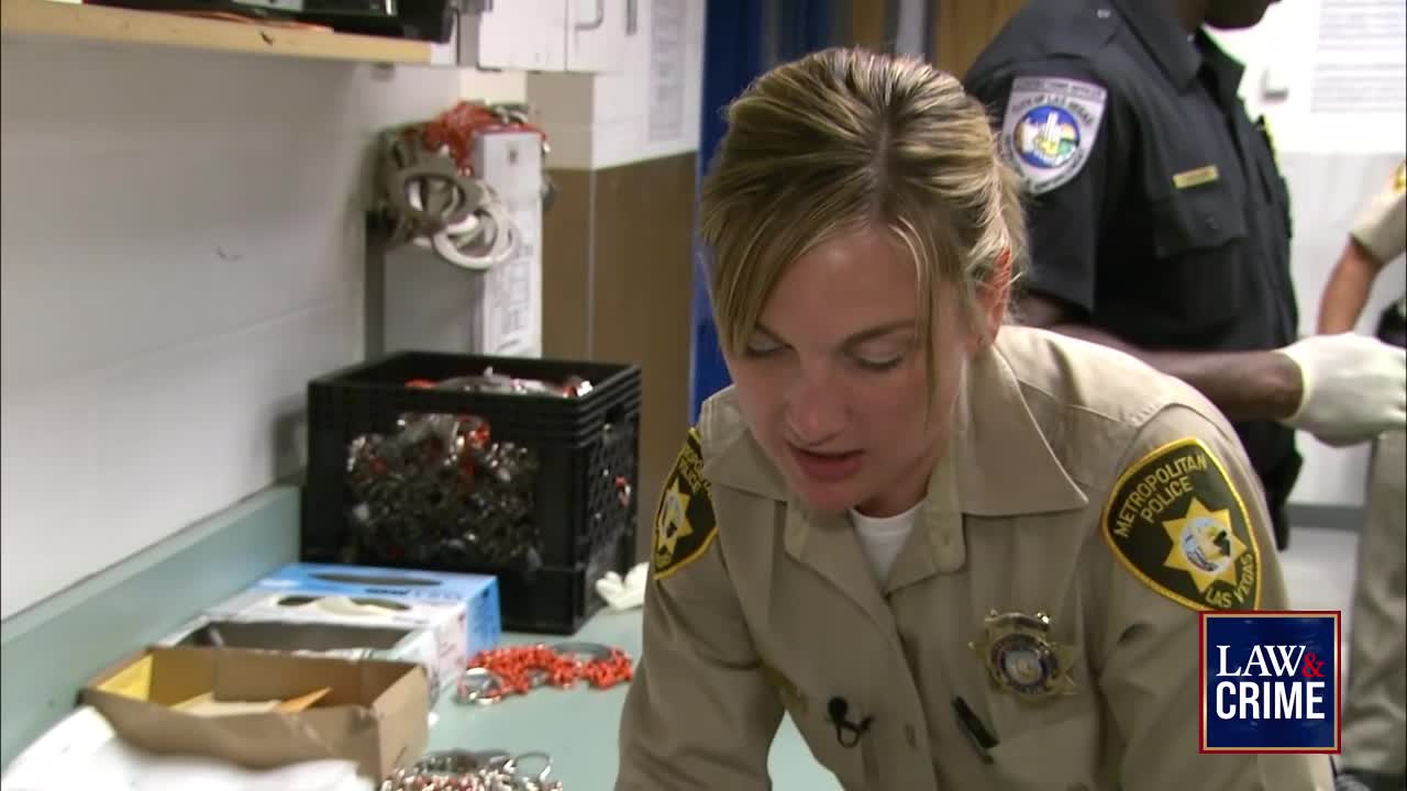 A woman in a Metropolitan Police uniform leans over a table, examining items. Another officer in uniform, wearing gloves, stands behind her, assisting with evidence.