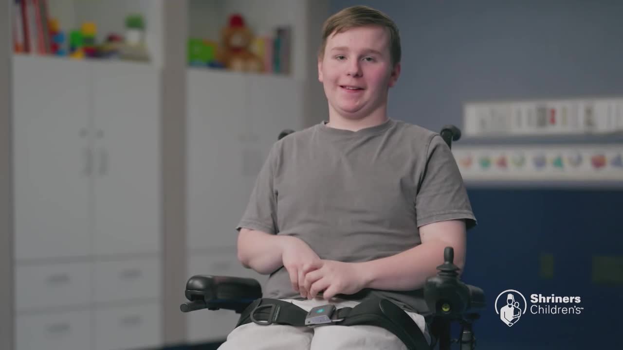 A young man sits in a powered wheelchair, looking directly ahead. He wears a simple gray t-shirt and appears to be in a room with children's toys visible in the background.
