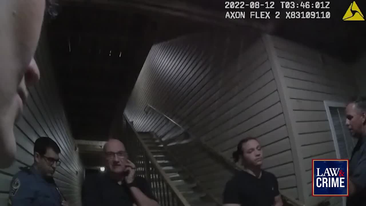 A man in glasses talks on his phone while two officers stand nearby. Another person, also in dark clothing, looks on as they stand outside an apartment building.