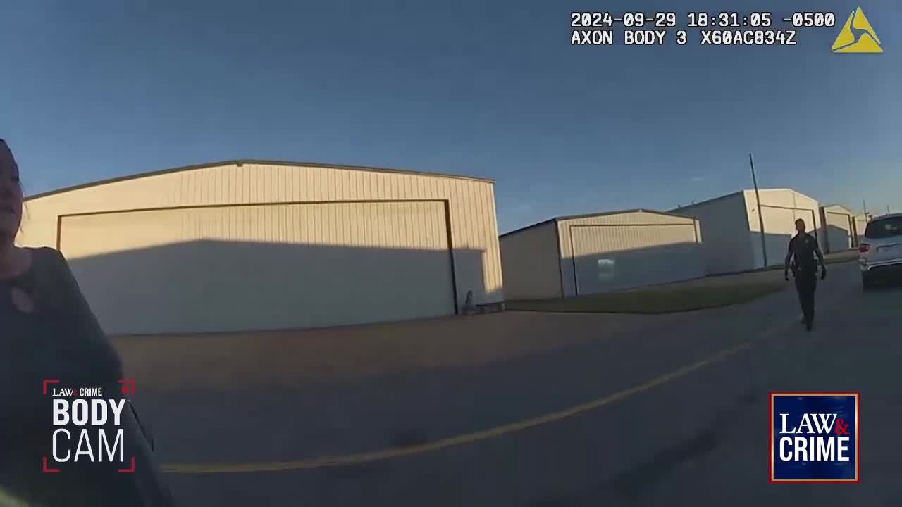 A police officer walks away from the camera toward a white SUV parked near hangars. The sun casts long shadows across the tarmac.