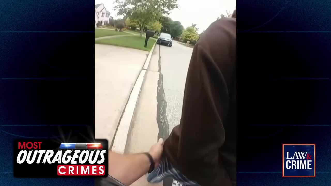 A dark sedan is stopped on the right side of a suburban street, its headlights off. A person in a brown hooded sweatshirt walks away from the camera, their hand held by another individual.