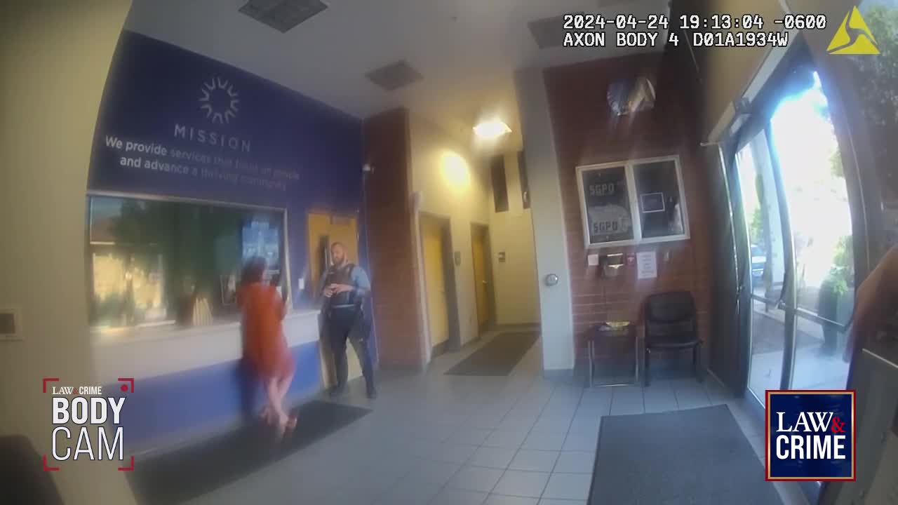A woman in an orange dress leans towards a counter, talking to an officer in uniform. The officer stands facing her, his hands behind his back.