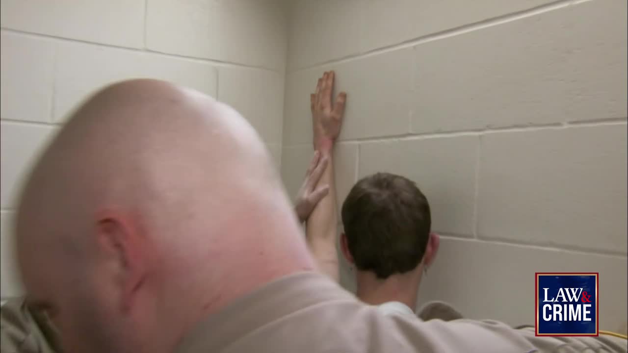 A man's arm is pressed against a cinder block wall, his hand reaching up. Another man, bald and in the foreground, is positioned behind him.