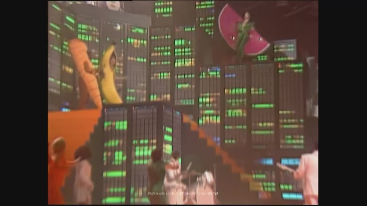 A person in a banana costume stands next to someone in a carrot costume on a raised platform. A woman in a green outfit with a pink fan-like prop stands on a higher level, overlooking a cityscape of green-lit panels.