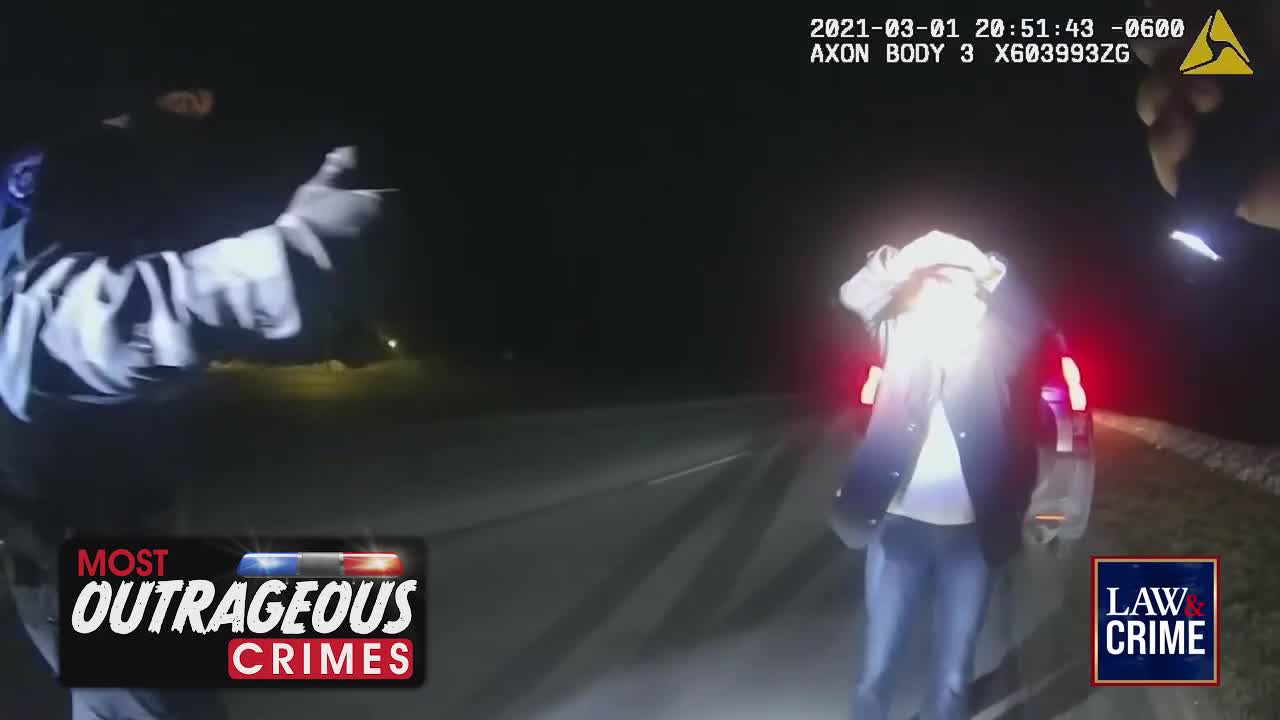 A person in jeans and a jacket stands with their hands over their head, illuminated by bright lights. An officer in a uniform with reflective stripes gestures with their left hand.