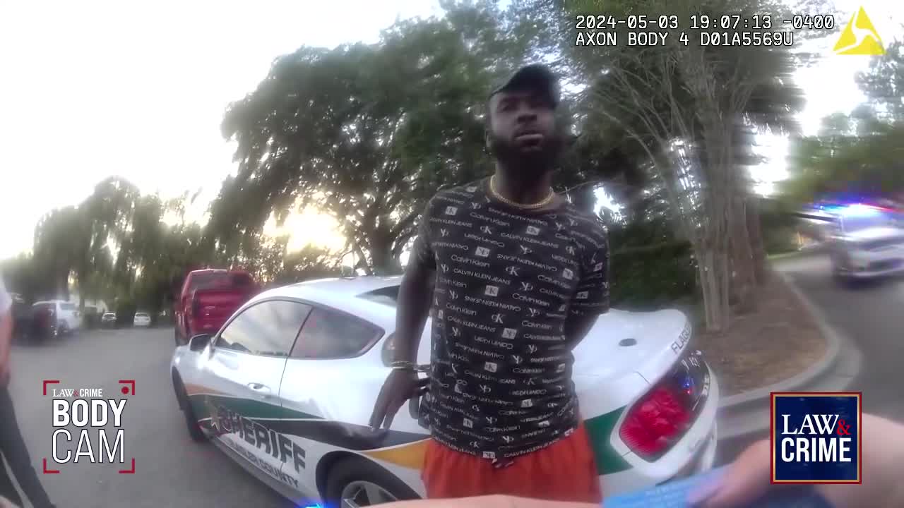 A man in handcuffs stands next to a sheriff's patrol car. Flashing blue and red lights from another vehicle illuminate the scene.