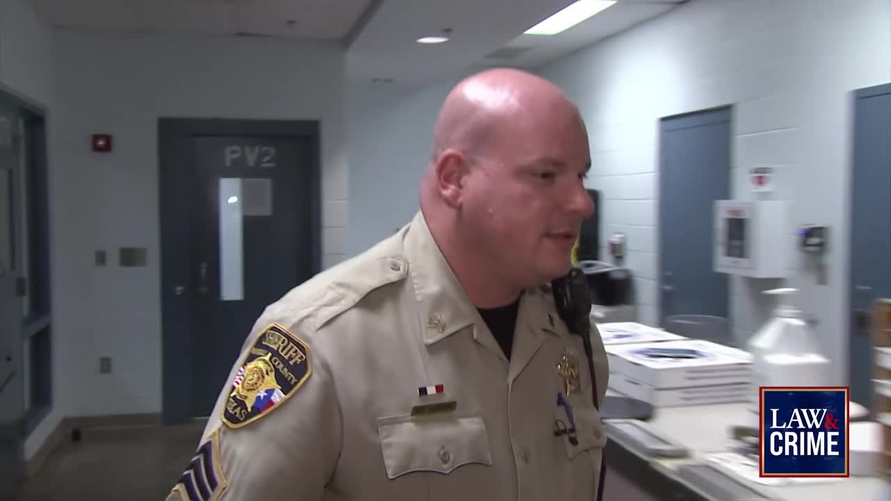 A sheriff's deputy in uniform walks past a cell door labeled "PV2." He's speaking, with a Law & Crime logo visible in the corner.
