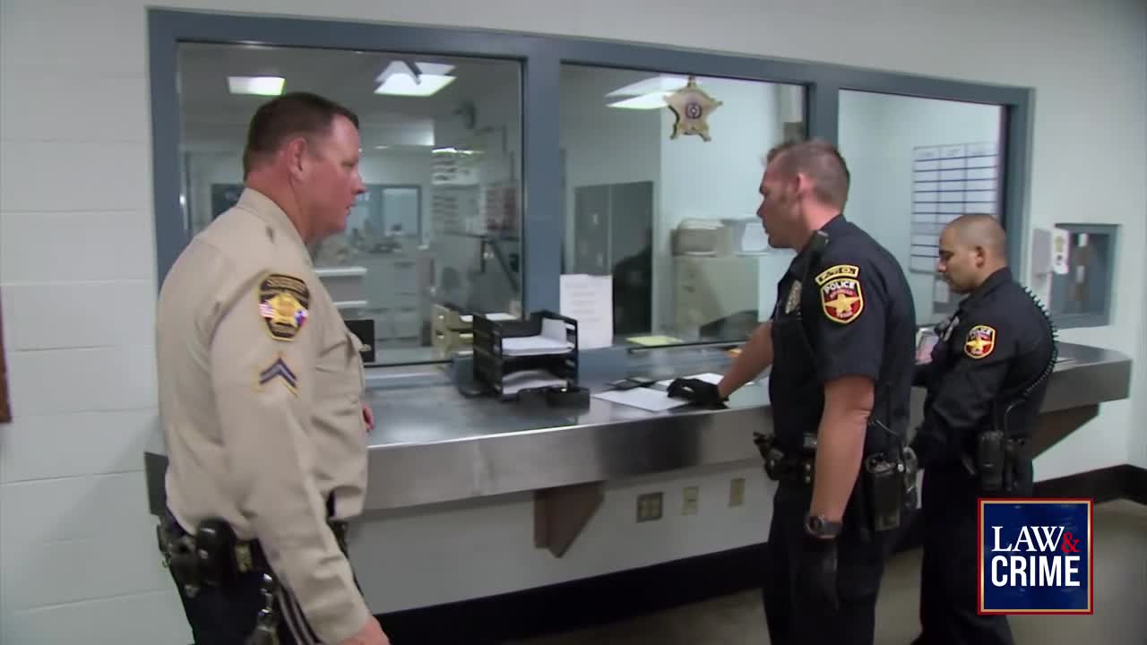 A sheriff's deputy in tan uniform talks to two officers in dark blue uniforms behind a counter. The officers appear to be in a secure facility, possibly a jail or courthouse, with a Law & Crime logo visible in the corner.