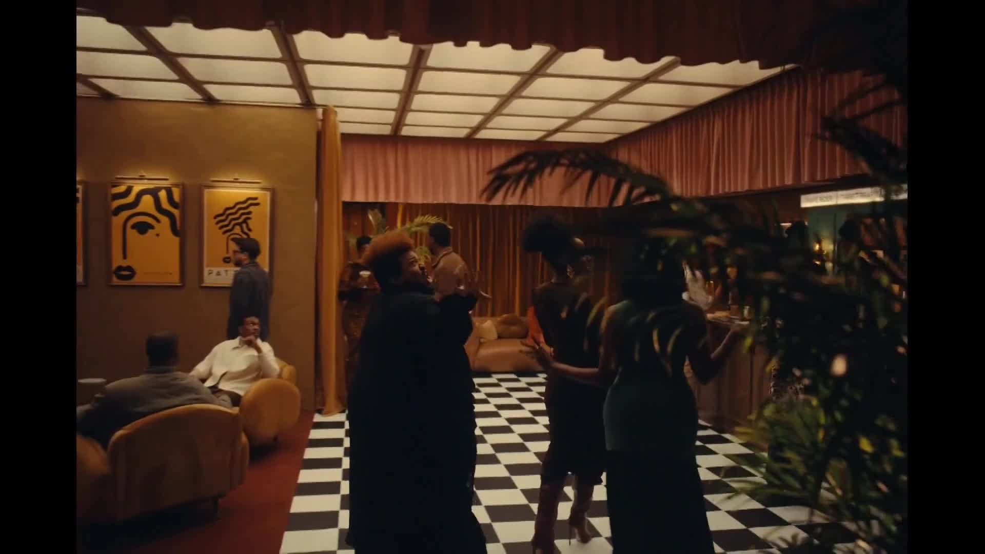 A group of people are gathered in a warmly lit room, some chatting while others dance on a checkered floor. A woman in a long black coat gestures as she speaks, while others in the background enjoy the party.
