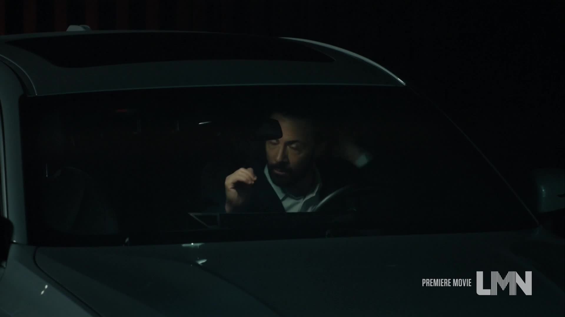 A man in a suit sits in the driver's seat of a car, fiddling with something in his lap. The scene is dark, and the LMN logo in the corner suggests this is a movie.
