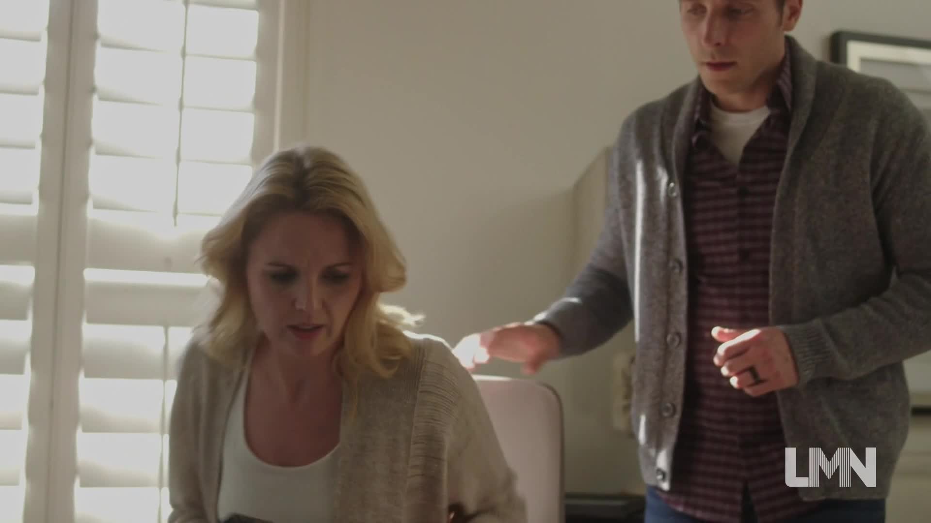 A woman in a cardigan looks down, her face etched with worry. A man, wearing a grey sweater, reaches out a hand toward her shoulder.
