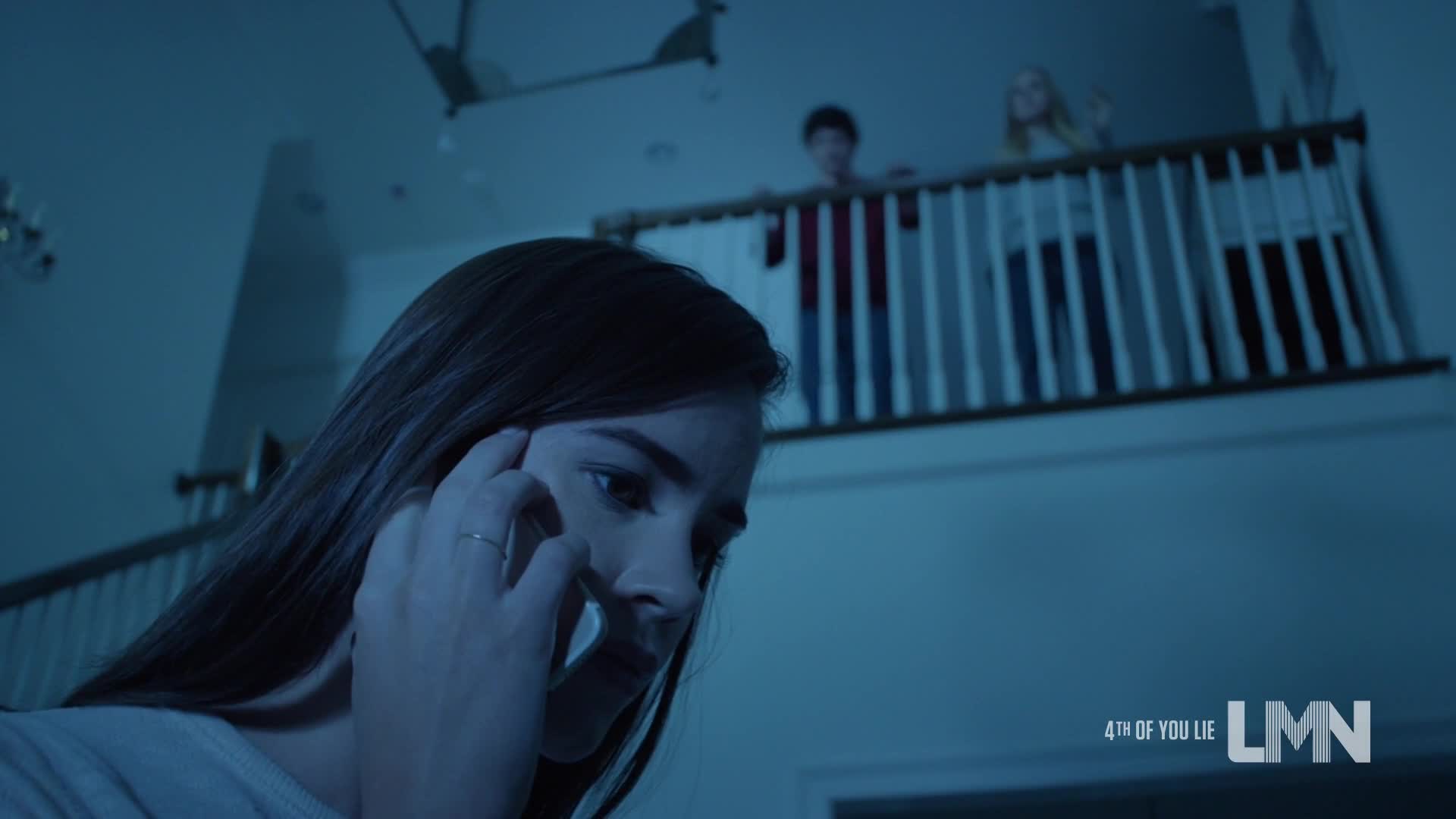 A woman, looking distressed, is on the phone, her hand pressed to her ear. Two figures stand on a balcony above, watching her.
