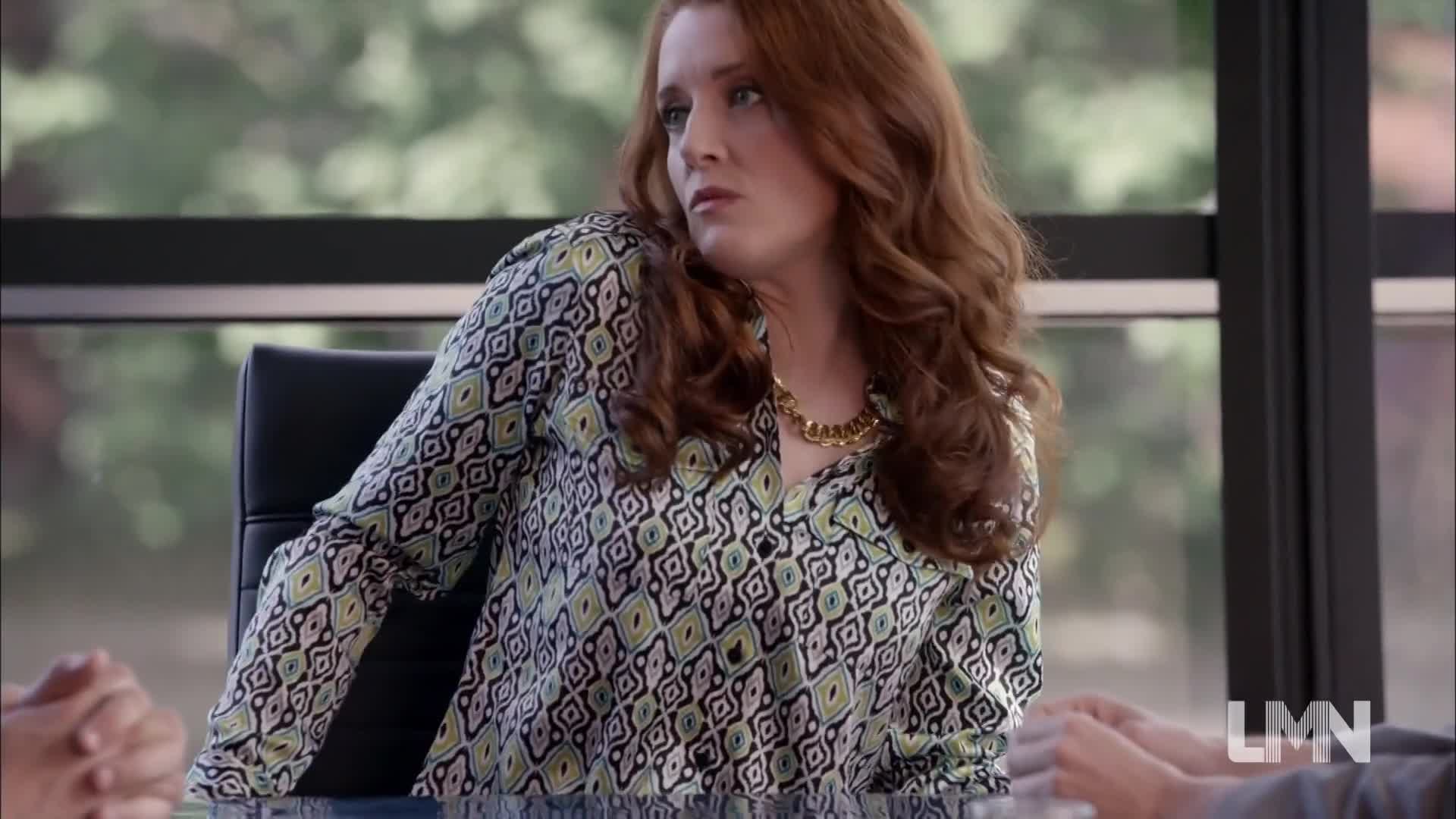 A woman with fiery red hair sits at a table, her eyes fixed on someone out of frame. She's wearing a patterned blouse and a gold necklace, leaning slightly to the side.
