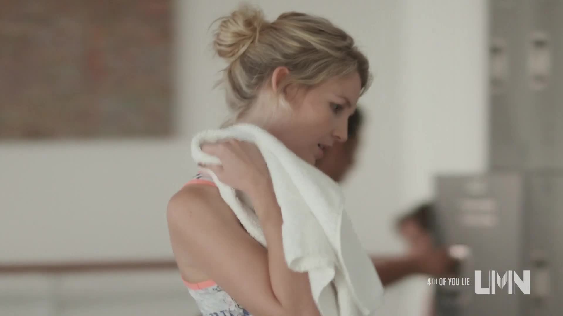 A woman with blonde hair in a bun wipes her neck with a white towel. She's in a gym, and there's someone else in the background.
