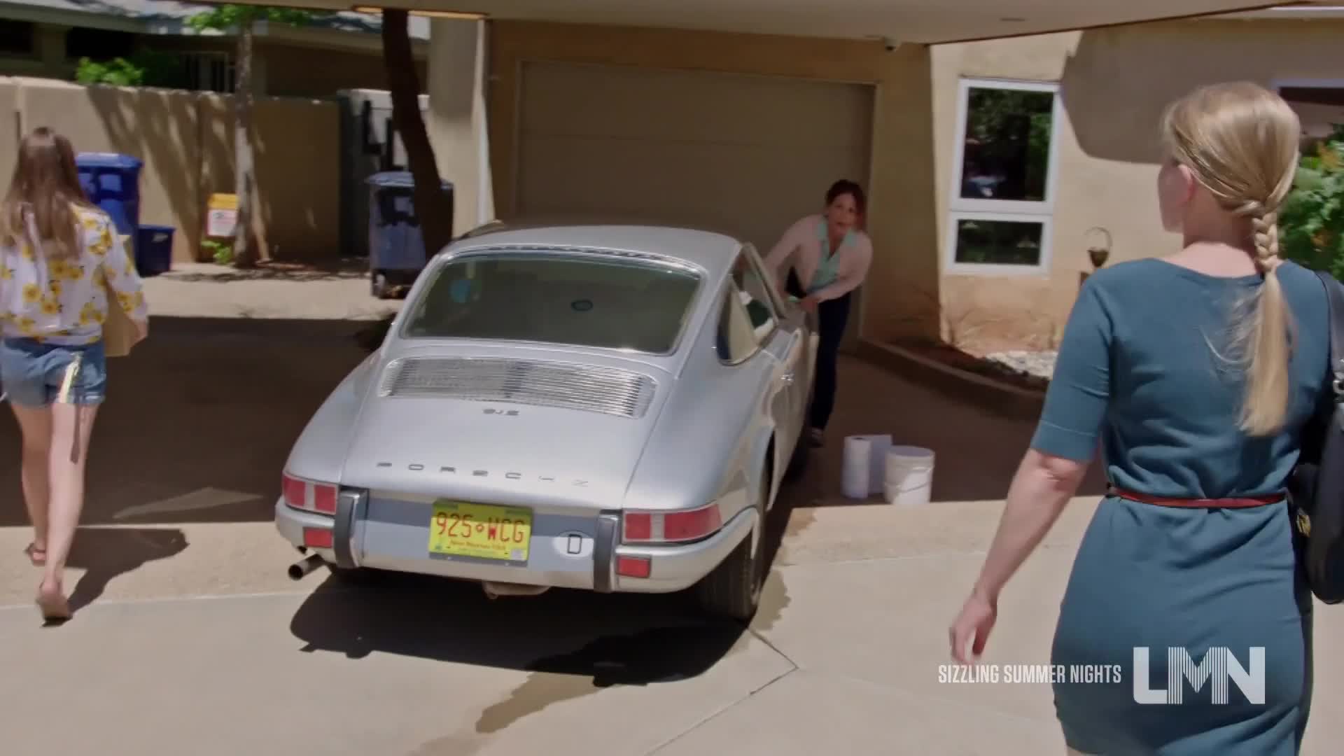 A woman in a light jacket leans into a silver Porsche parked in a driveway. Another woman walks past, heading away from the car, while a third woman approaches.
