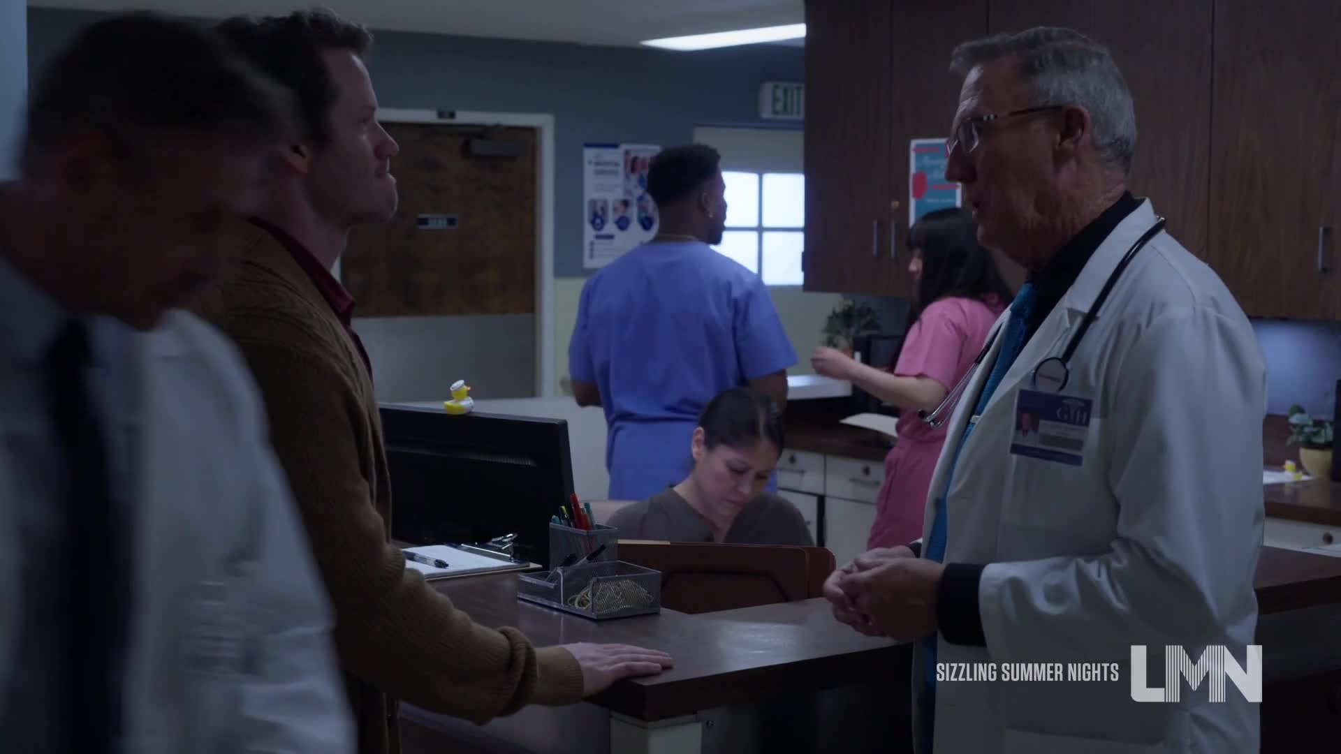 A doctor in a white coat gestures while speaking to two men in a hospital reception area. Behind them, a nurse in pink and a man in blue scrub tops are busy at a counter.
