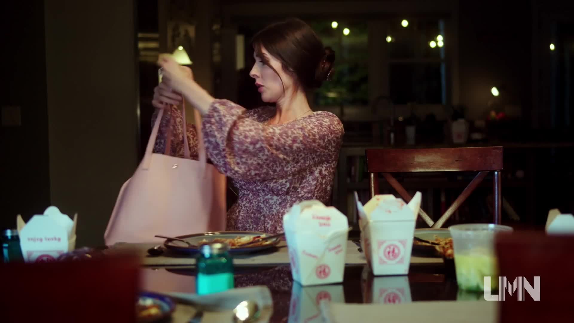 A woman in a floral dress pulls items from a large pink tote bag at a dining table set for a meal. Chinese takeout containers and a glass of green liquid sit nearby.
