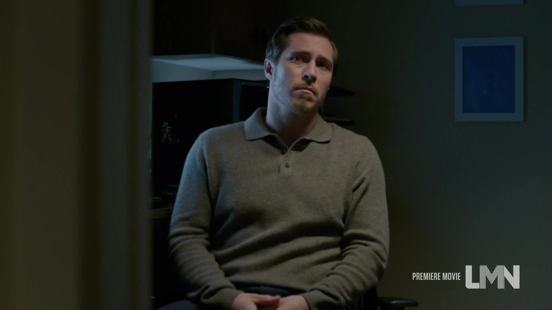 A man in a gray sweater sits in a chair, looking off to the side with a serious expression. The room is dimly lit, and a Lifetime Movie logo is visible in the corner.

