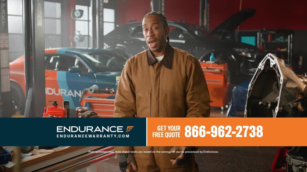 A man in a brown work jacket stands in an auto repair shop, speaking directly to the camera. Behind him, a car with a blue and orange design is parked on a lift.
