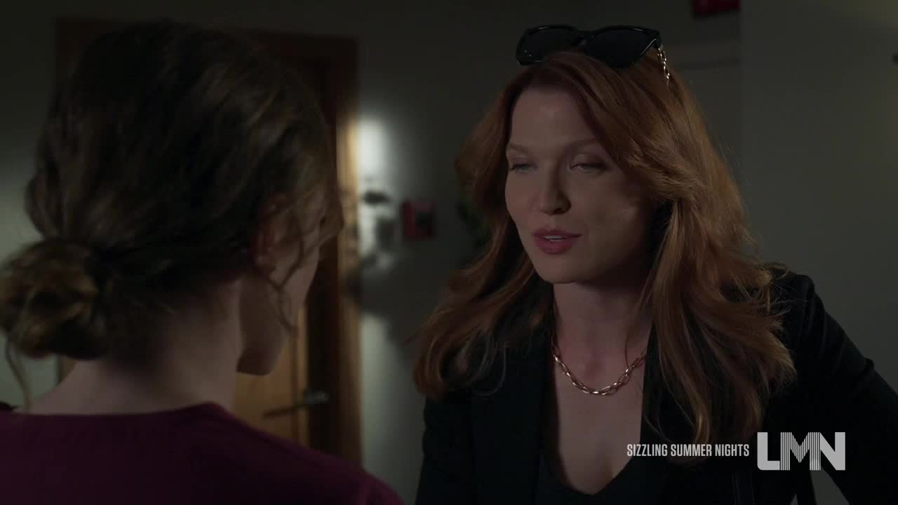 A woman with red hair is speaking, her eyes focused on someone out of frame. Sunglasses rest atop her head, and a delicate necklace catches the light.
